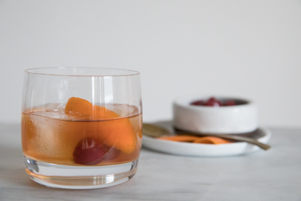 Low ball cocktail glass on marble counter. Large ice cubes in the glass with cherries and orange peel garnishing a Bourbon and Honey Old Fashioned Cocktail. Additional Garnishes in the background.