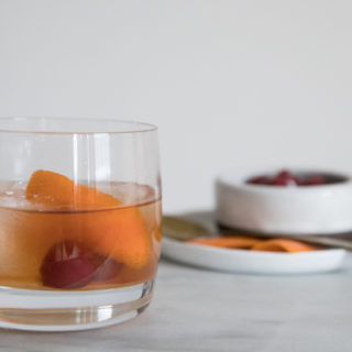 Low ball cocktail glass on marble counter. Large ice cubes in the glass with cherries and orange peel garnishing a Bourbon and Honey Old Fashioned Cocktail. Additional Garnishes in the background.