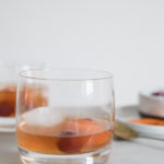 Low ball cocktail glass on marble counter. Large ice cubes in the glass with cherries and orange peel garnishing a Bourbon and Honey Old Fashioned Cocktail. Additional Garnishes in the background.