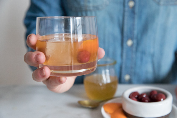 Low ball cocktail glass in a hand. Large ice cubes in the glass with cherries and orange peel garnishing a Bourbon and Honey Old Fashioned Cocktail. Additional Garnishes in the background on a marble countertop.